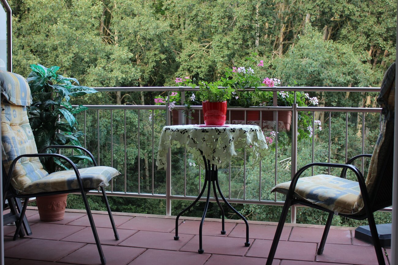 Der Balkon ist mit zwei Stühlen und einem kleinen Tisch ausgestattet. Auf dem Tisch steht ein Blumentopf mit einer grünen Pflanze. Im Hintergrund sind Bäume und Blumen zu sehen, die dem Ort eine ruhige und einladende Atmosphäre verleihen.