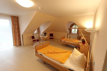 Das Bild zeigt ein helles Zimmer mit zwei Pflegebetten, die mit einer orangefarbenen Decke bedeckt sind. Neben den Betten befindet sich ein Tisch mit Stühlen und einer Pflanze. Ein Fenster lässt natürliches Licht herein, und die Wände sind in sanften Farben gehalten.